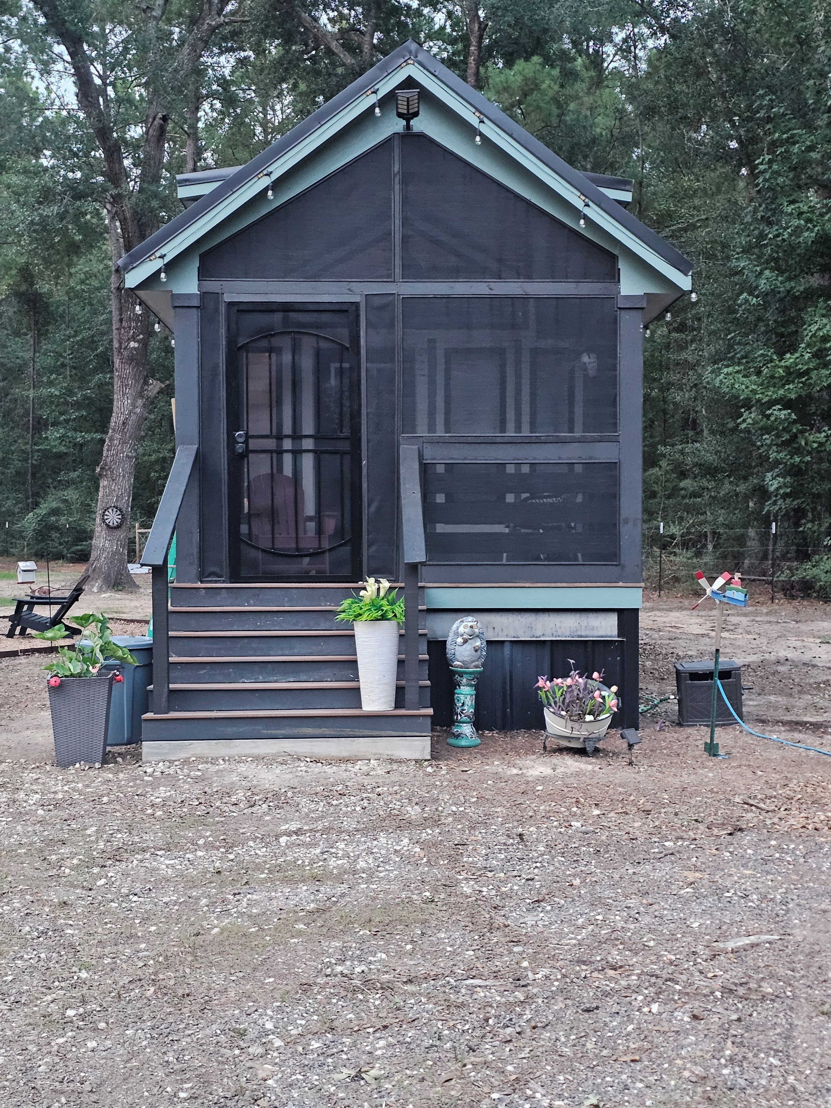 A small, dark-colored cabin with a porch, steps leading up to the entrance, and potted plants around the base.