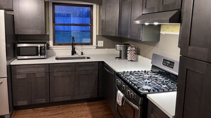 A modern kitchen with dark cabinetry, a stainless steel microwave, a gas stove, and a window with a view of trees. - Entire Residential home (Darien)