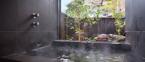 A modern spa with a hot tub, dark tiled walls, and a view of a wooden fence and trees.
