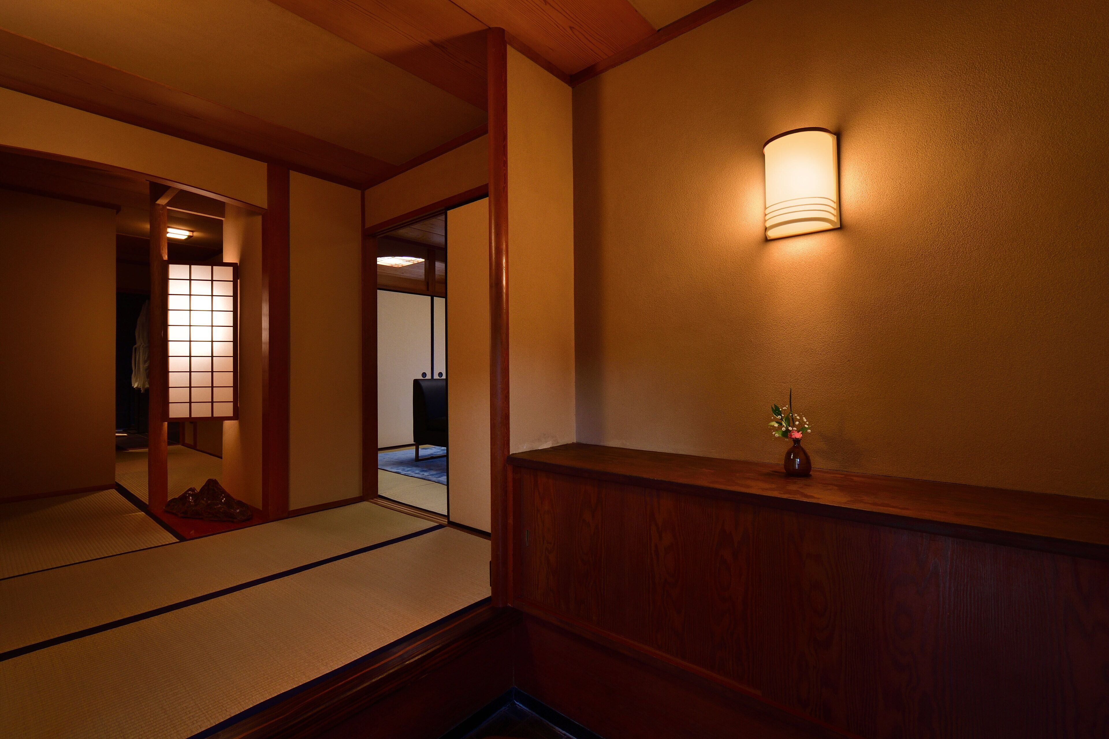 Un interior japonés tradicional con suelo de tatami, un banco de madera, un aplique y un pequeño jarrón con flores.