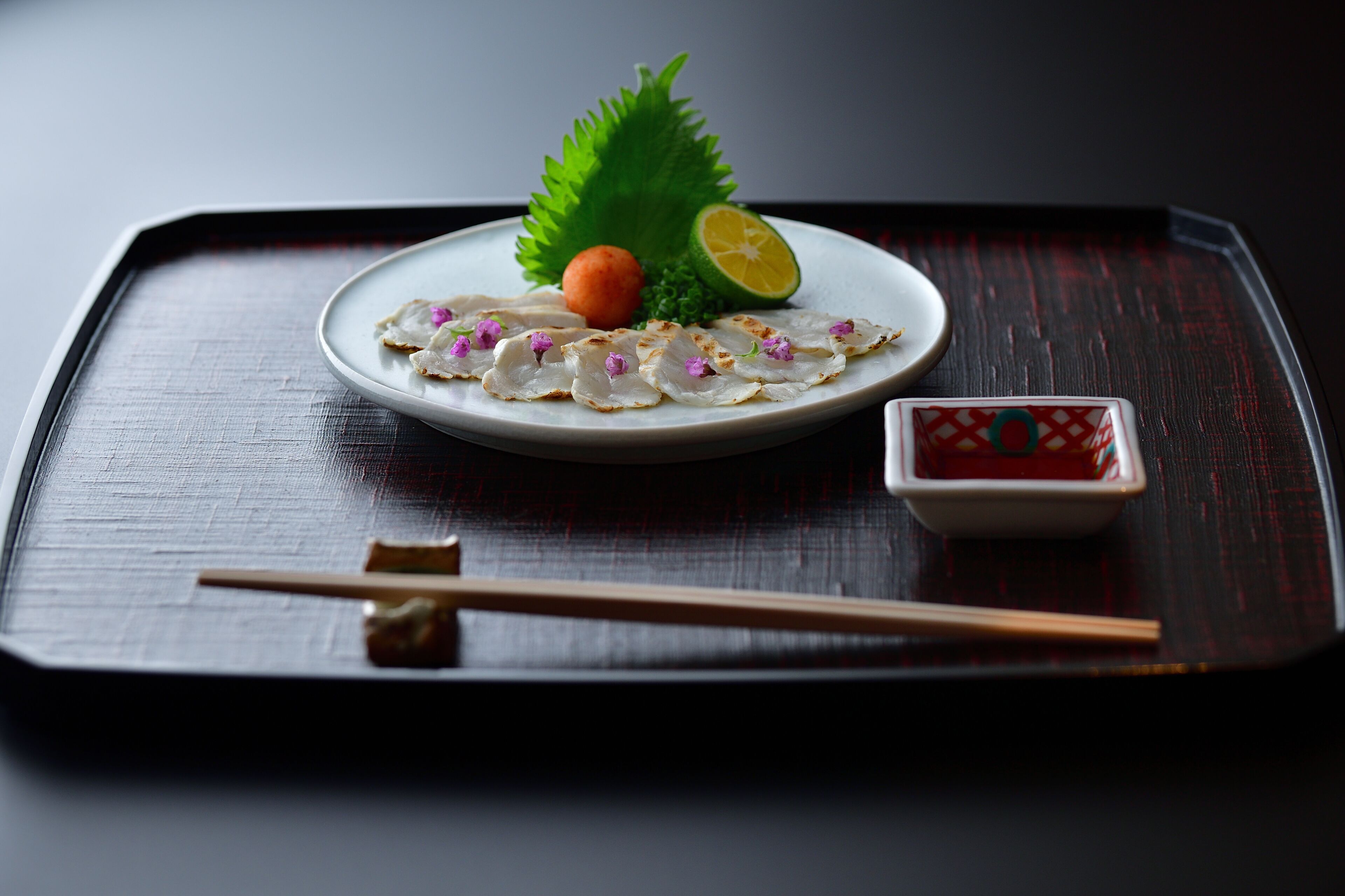 A plate of sashimi with a slice of lemon, a small orange ball, and a decorative leaf on a dark tray.