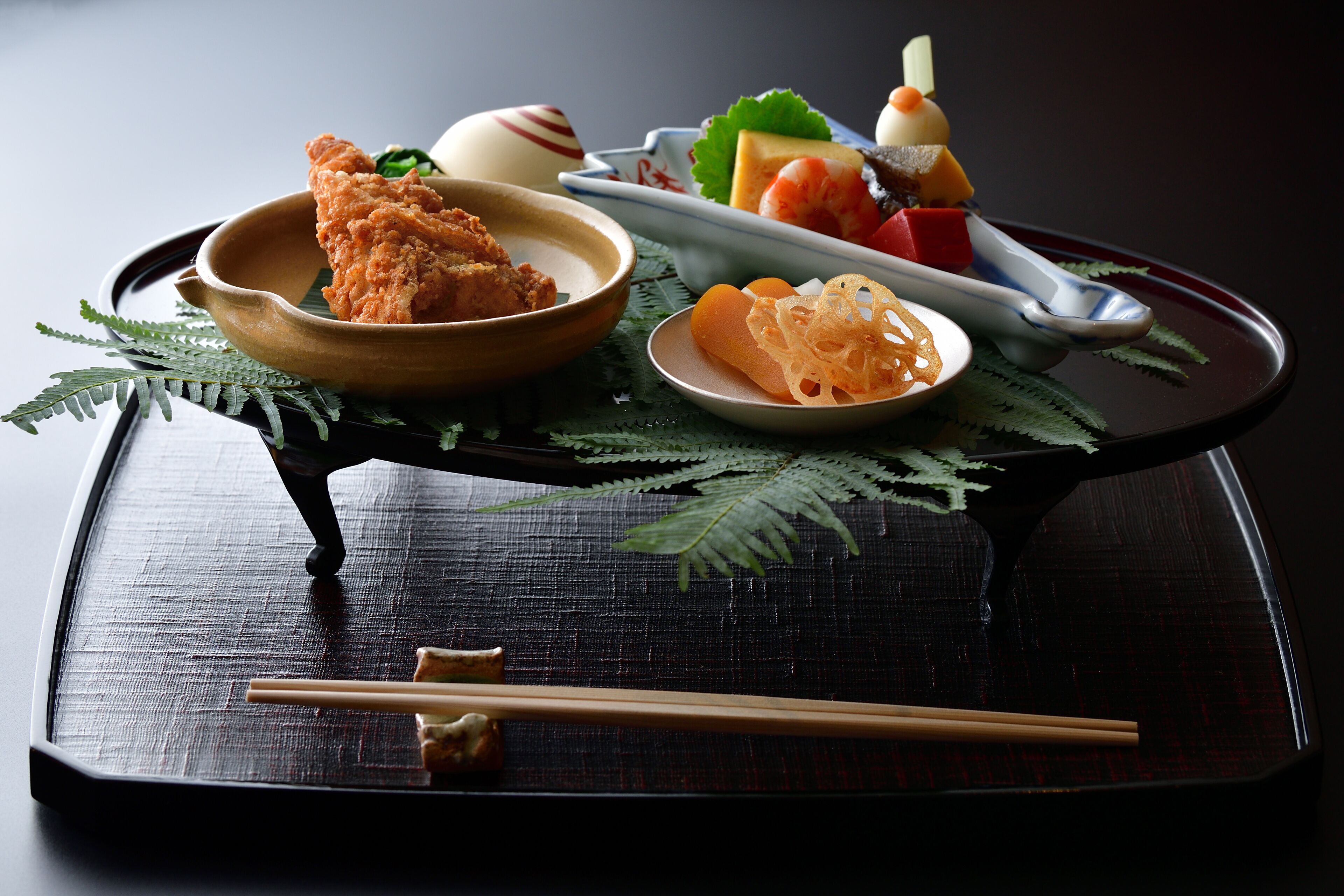 Un repas japonais traditionnel composé de divers plats, notamment du poulet frit, des sashimis et des légumes marinés, servi sur un plateau en bois avec des baguettes et une cuillère.