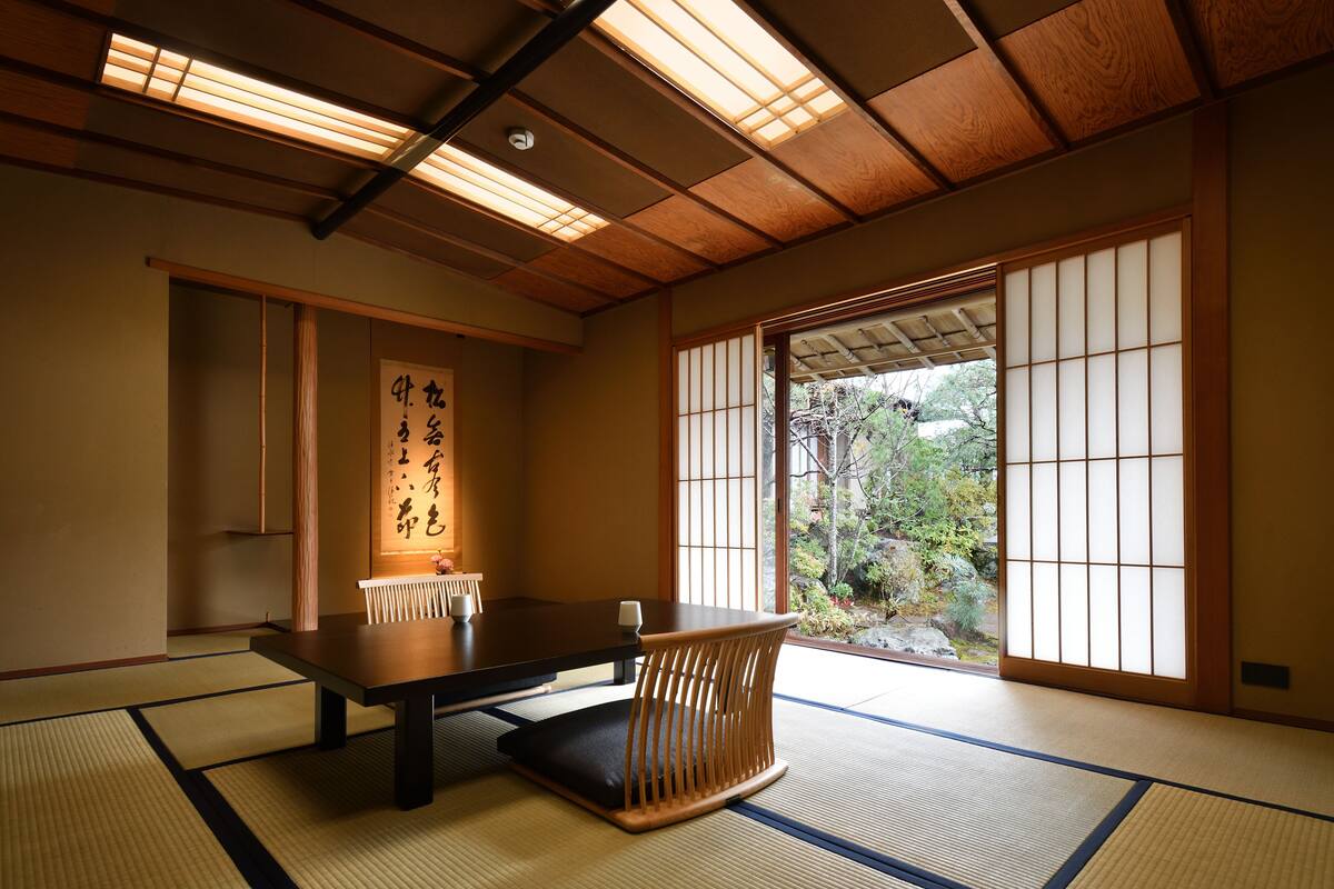 A traditional Japanese room with tatami flooring, a low table with cushions, sliding doors, and a view of greenery outside.