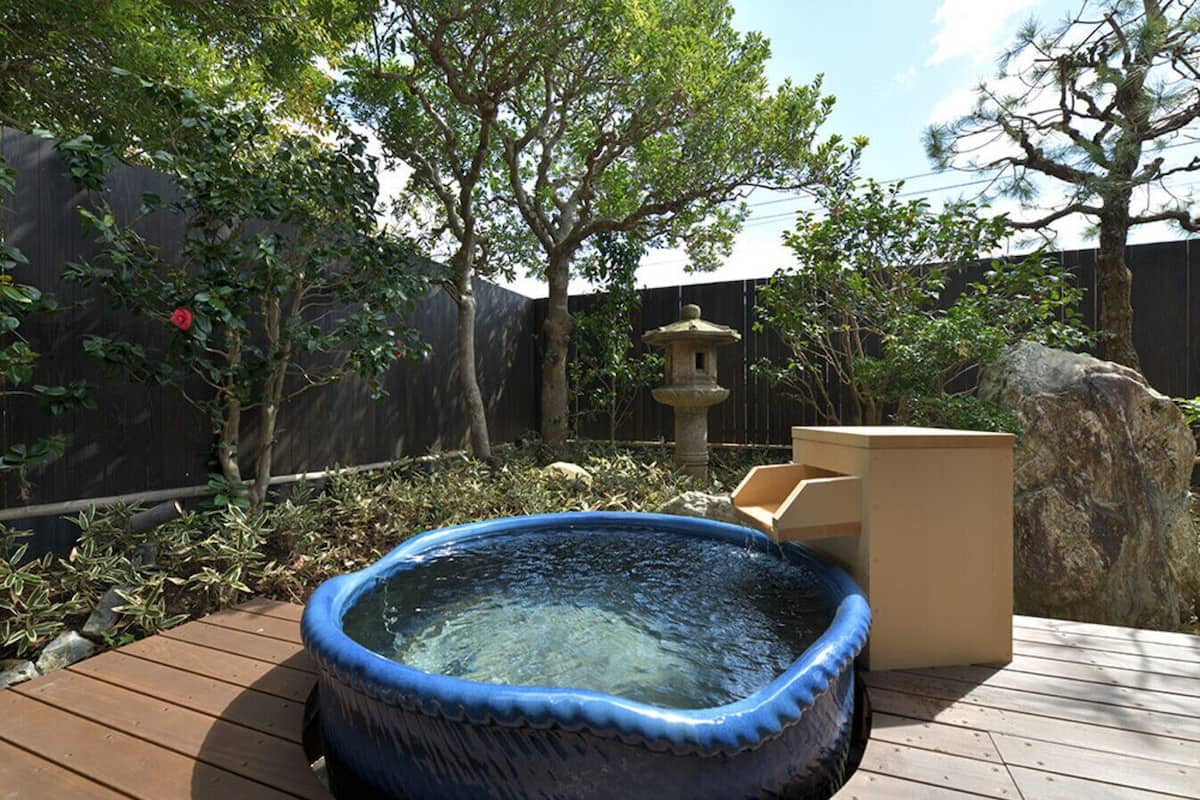 A hot tub on a wooden deck with a stone fountain and a wooden structure in the background.