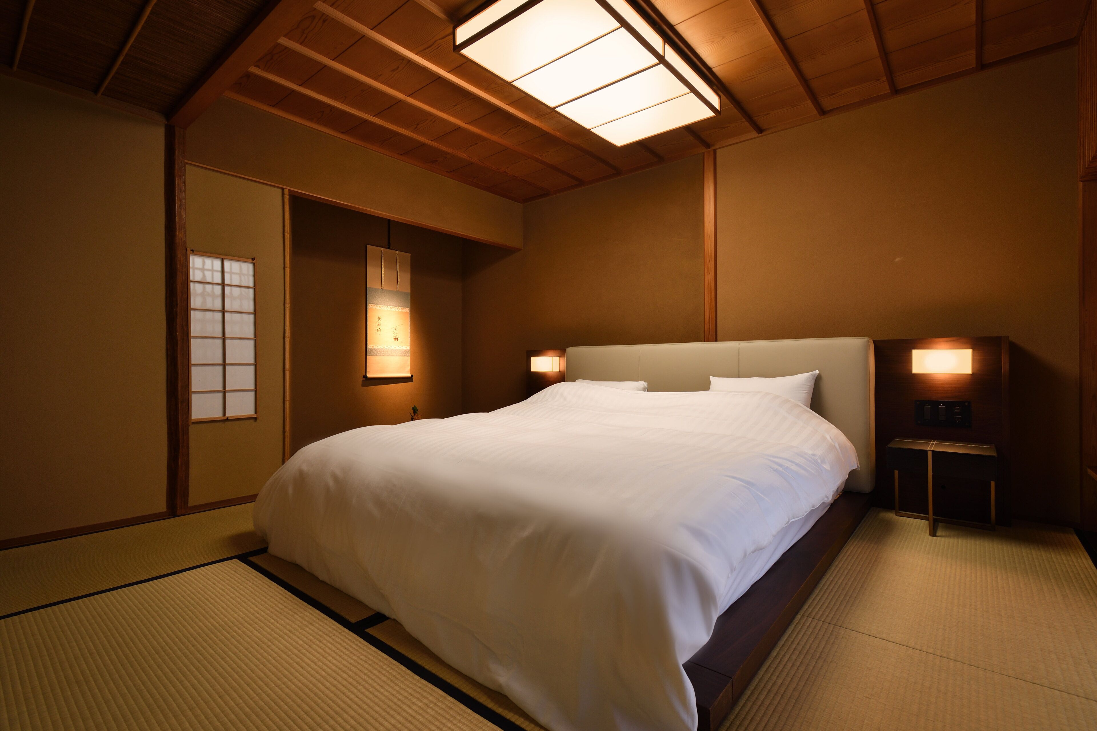 A traditional Japanese-style bedroom with tatami flooring, a large bed with white bedding, and wooden beams on the ceiling.