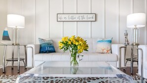 A modern living room with a white couch, a glass coffee table, and a vase of yellow flowers. There are two lamps on either side of the couch, and a wall-mounted sign that reads "life is better at the beach." - Gulf Front PCB Condo, Stunning Views, Pool, Balcony, Sleeps 6 (Panama City Beach)