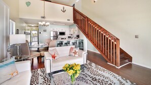 A spacious living area with a white sofa, a coffee table with a vase of flowers, a wooden staircase, and a kitchen in the background. - Gulf Front PCB Condo, Stunning Views, Pool, Balcony, Sleeps 6 (Panama City Beach)