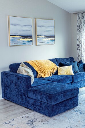 A blue velvet sofa with a yellow throw blanket, two framed artworks on the wall, and a patterned rug.