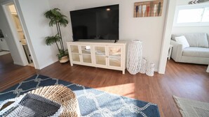 Ein Wohnzimmer mit einem Flachbildfernseher auf einem weißen Schrank, einer großen Topfpflanze und einem blauen Teppich mit Muster.