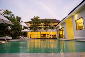 A swimming pool with lounge chairs and umbrellas, surrounded by lush greenery and a modern building with large windows.