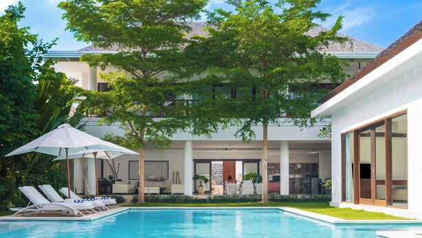 Un’area piscina moderna con lettini e ombrellone, immersa nel verde e affacciata su un edificio a più piani con ampie finestre.