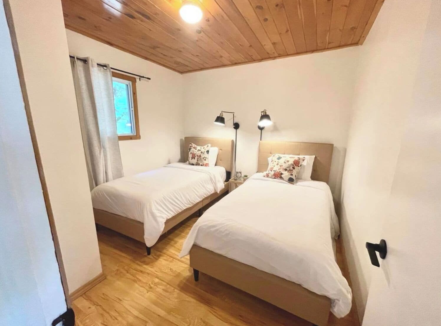 A room with two single beds, wooden flooring, a window with curtains, and a ceiling light.
