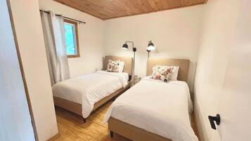 A room with two single beds, wooden flooring, a window with curtains, and a ceiling light.