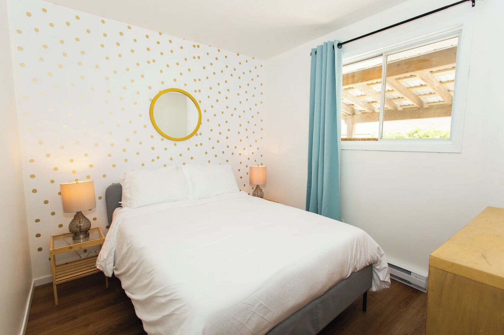 A bedroom with a bed, two bedside tables with lamps, a round mirror, a window with curtains, and a wooden headboard.