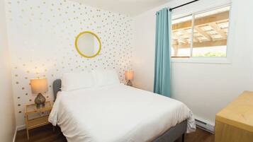 A bedroom with a bed, two bedside tables with lamps, a round mirror, a window with curtains, and a wooden headboard.