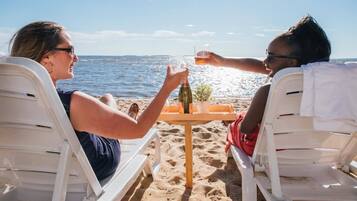 Twee personen zitten op witte strandstoelen aan een houten tafel met drankjes en een klein plantje, met uitzicht op de zee.