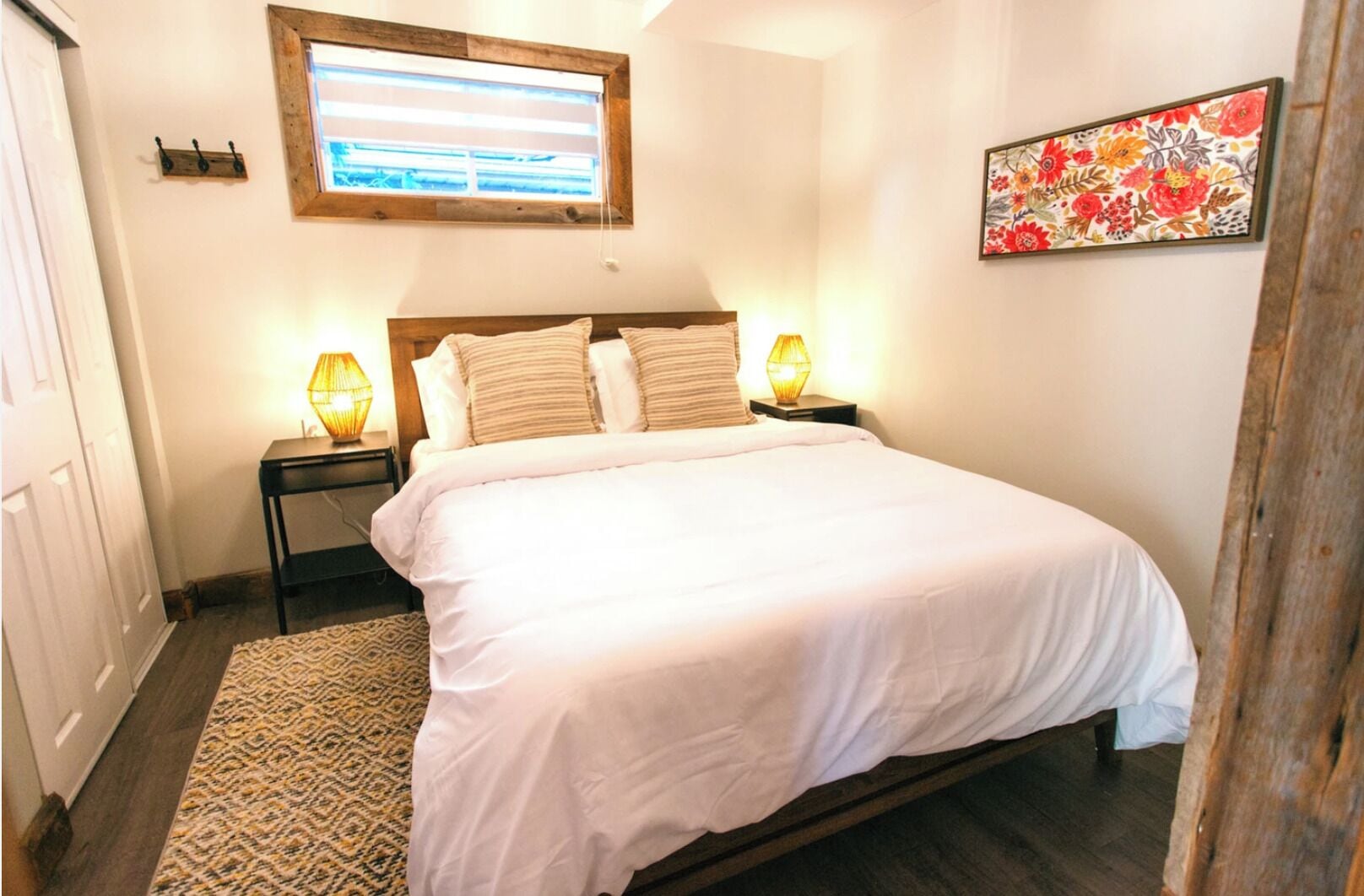 Une chambre avec un grand lit, deux lampes de chevet, une fenêtre avec des stores, une œuvre d’art florale accrochée au mur et un tapis à motifs.