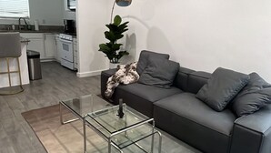 Ein modernes Wohnzimmer mit einem grauen Sofa, einem Glastisch und einer Küche im Hintergrund.