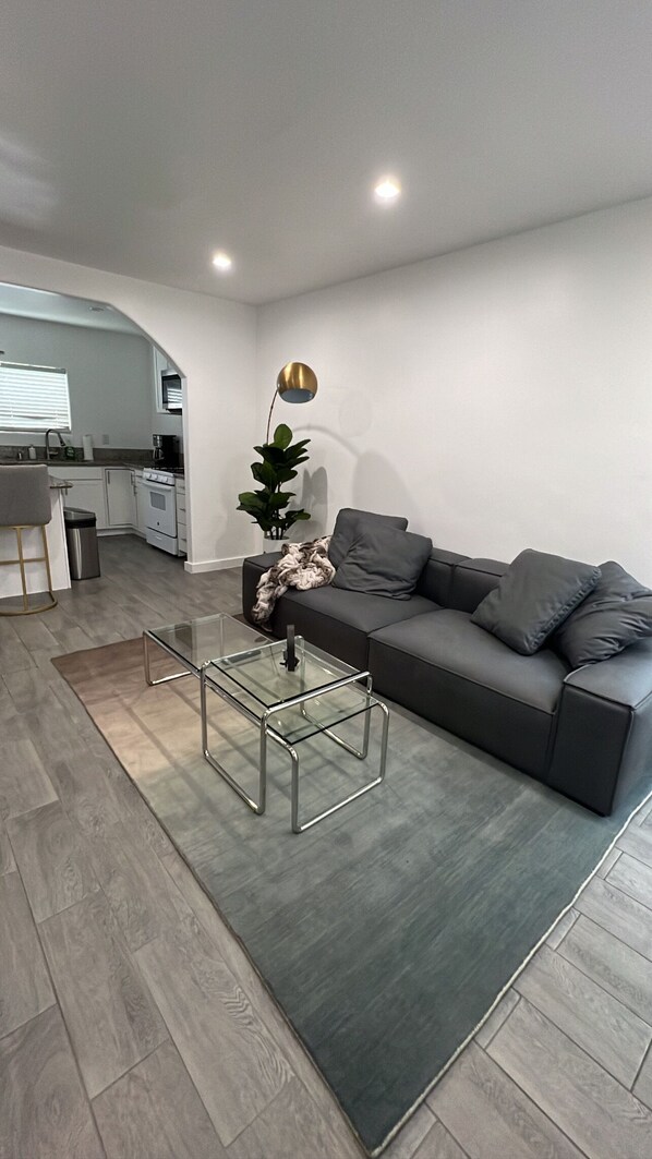 A modern living room with a grey sofa, a glass coffee table, and a kitchen in the background.