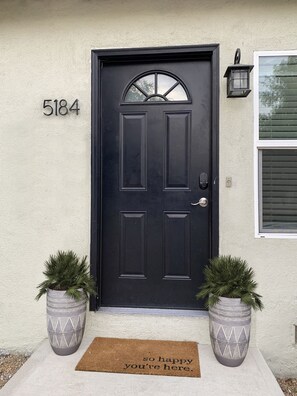 A black front door with the number 5184, a doormat with a welcoming message, and two potted plants on either side.