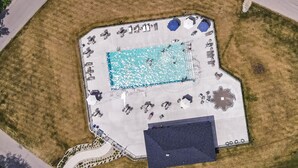 Vista aérea de una zona de piscina con una piscina rectangular, sillones de descanso alrededor y un área de estar cubierta.