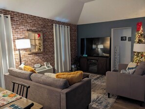 A modern living room with a brick wall, a flat-screen TV, two sofas, a fireplace, and a Christmas tree.