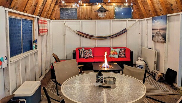 A cozy, rustic interior with a wooden ceiling, a red sofa, a round table with a fire, and a hammock.