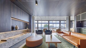 Una sala de estar moderna con un sofá de cuero, un otoman redondo y una mesa de centro. Hay dos sillones y una estantería con objetos decorativos. Ventanas amplias ofrecen vistas al exterior.