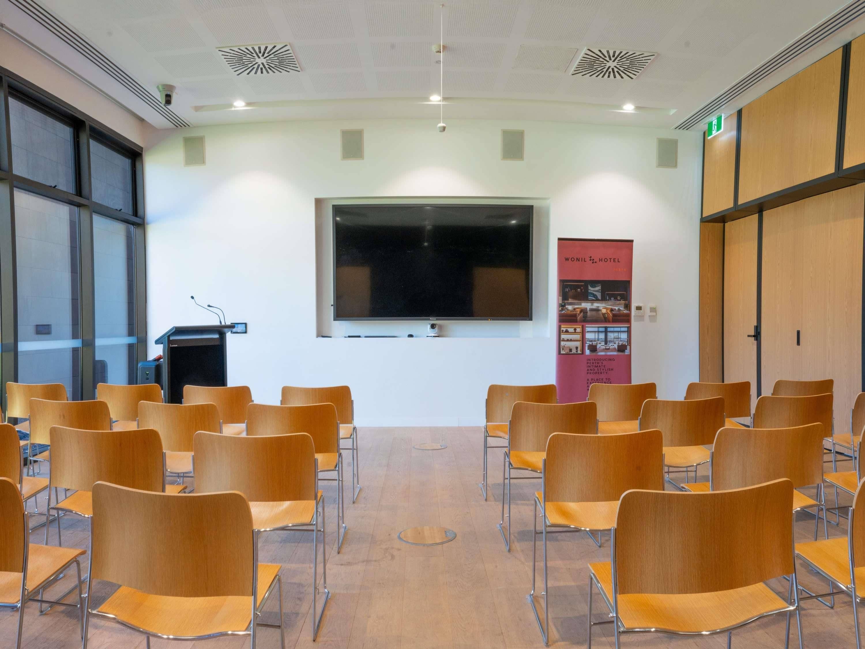 Un salón de conferencias con filas de sillas, una pantalla grande, un atril y un cartel en la pared.