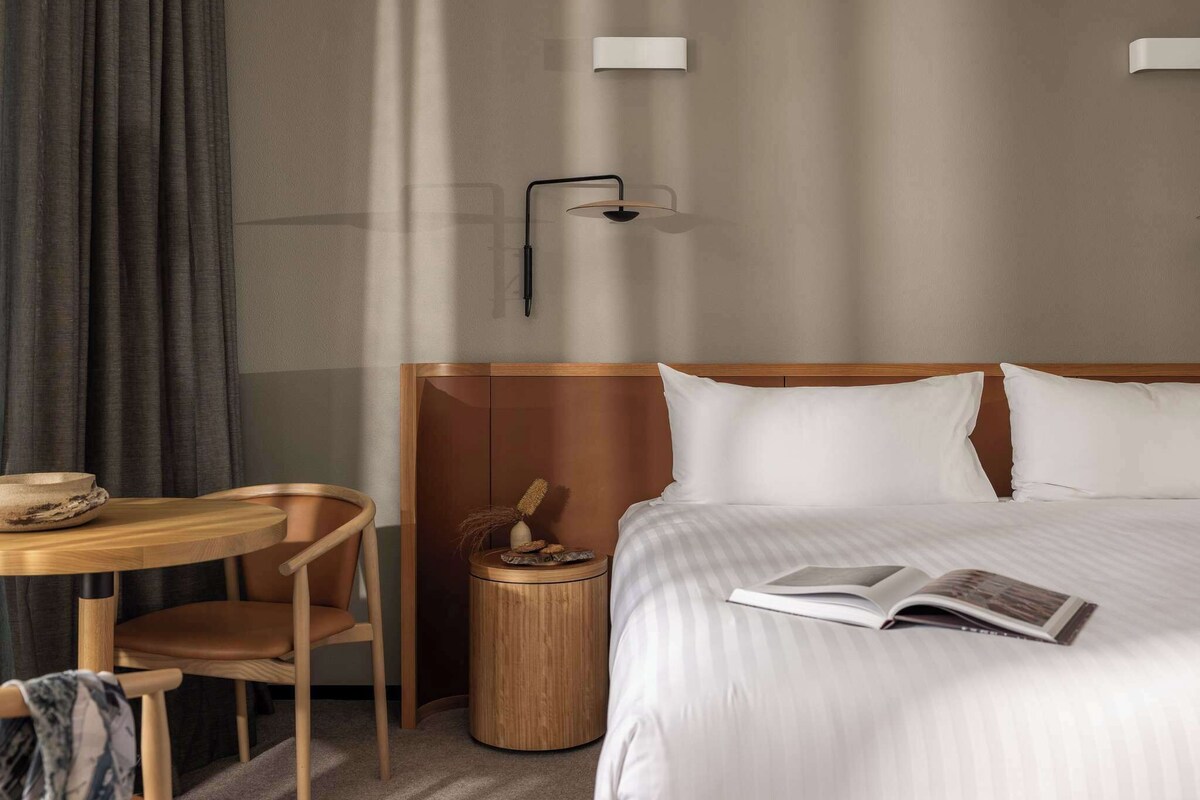 Ein Hotelzimmer mit einem Bett, einem Nachttisch, einem Sessel, einer Lampe und einer Wandleuchte.
