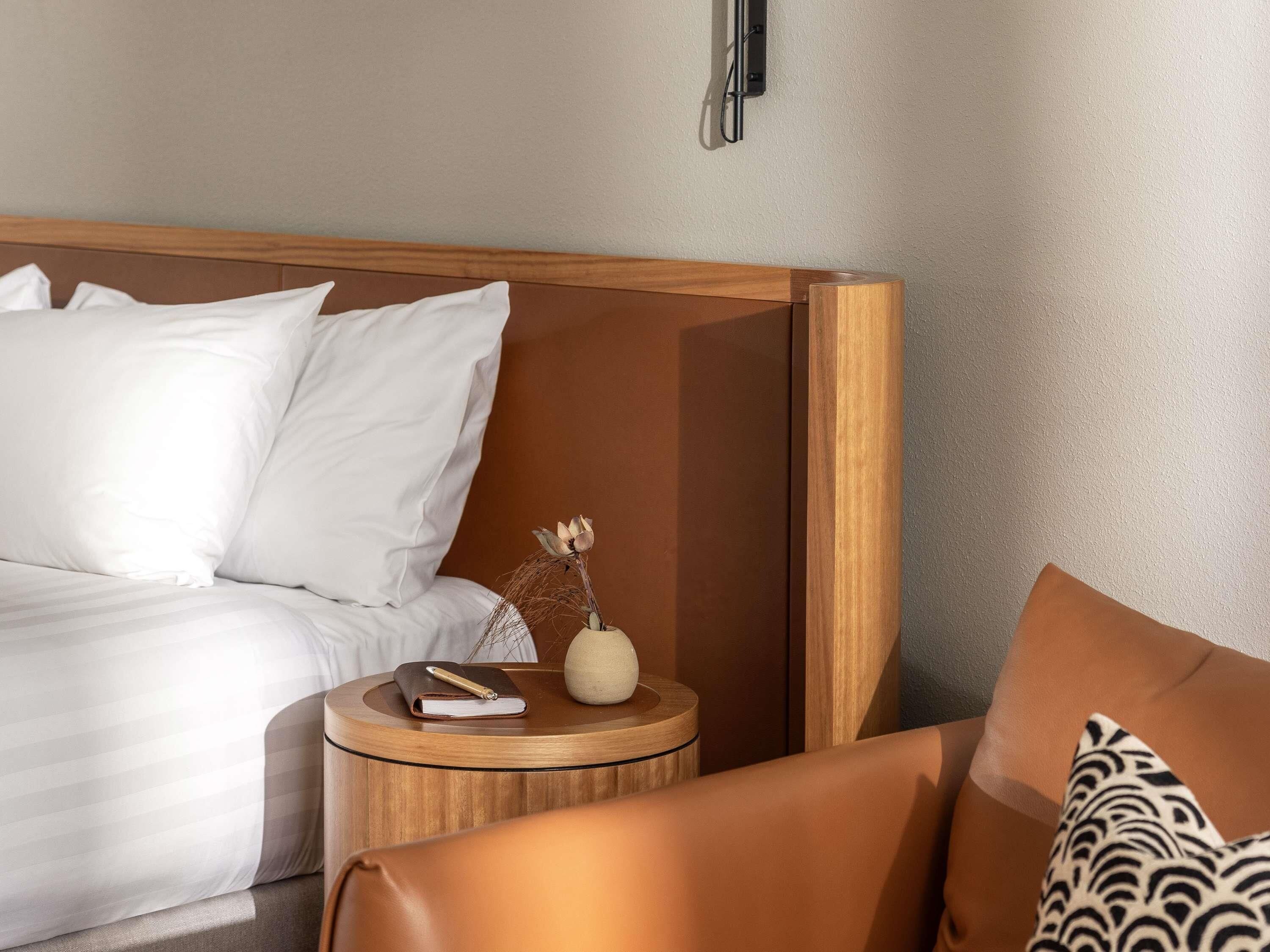Una cama bien hecha con sábanas blancas, una mesita de noche de madera con un libro y un florero, y un sillón de cuero marrón con un cojín estampado.