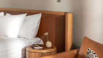 Una cama bien hecha con sábanas blancas, una mesita de noche de madera con un libro y un florero, y un sillón de cuero marrón con un cojín estampado.