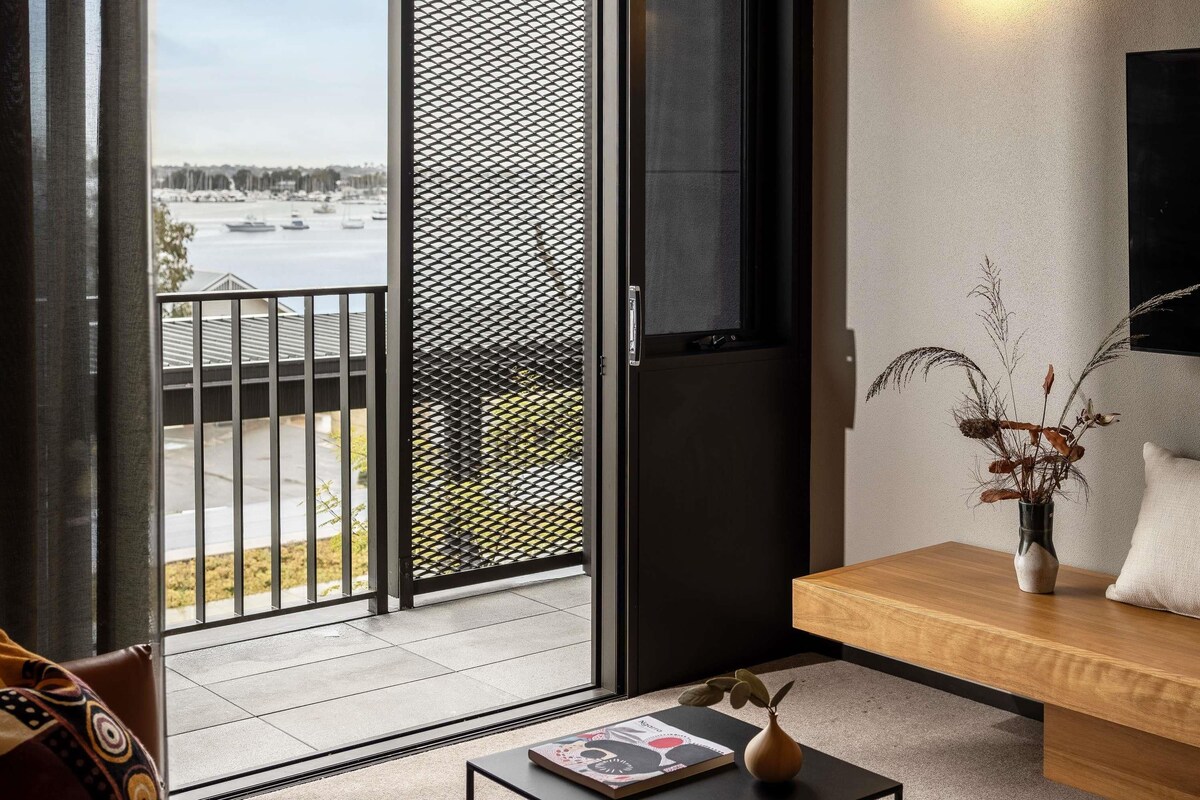 A modern living room with a sliding glass door leading to a balcony, a wooden bench, a small table with a book, and a vase with dried plants.