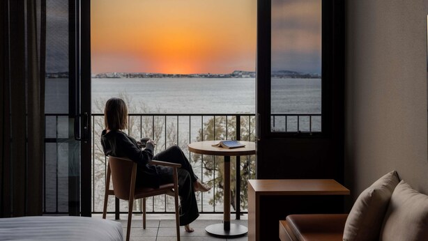 A room with a view of a sunset over water, a person sitting at a table, and a bed in the foreground. - Wonil Hotel Perth - Handwritten Collection (Crawley)