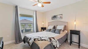 Una camera da letto con un letto, una ventola a soffitto, un comodino, una lampada e una finestra con tende.