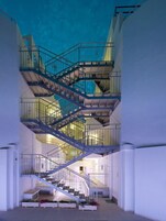 Un interior de varios niveles con una escalera central, paredes blancas y una barandilla azul.