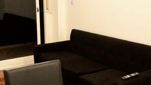 Ett vardagsrum med en svart soffa, ett glasbord täckt med plast och en skjutdörr i glas som leder ut till ett mörkt område utomhus.