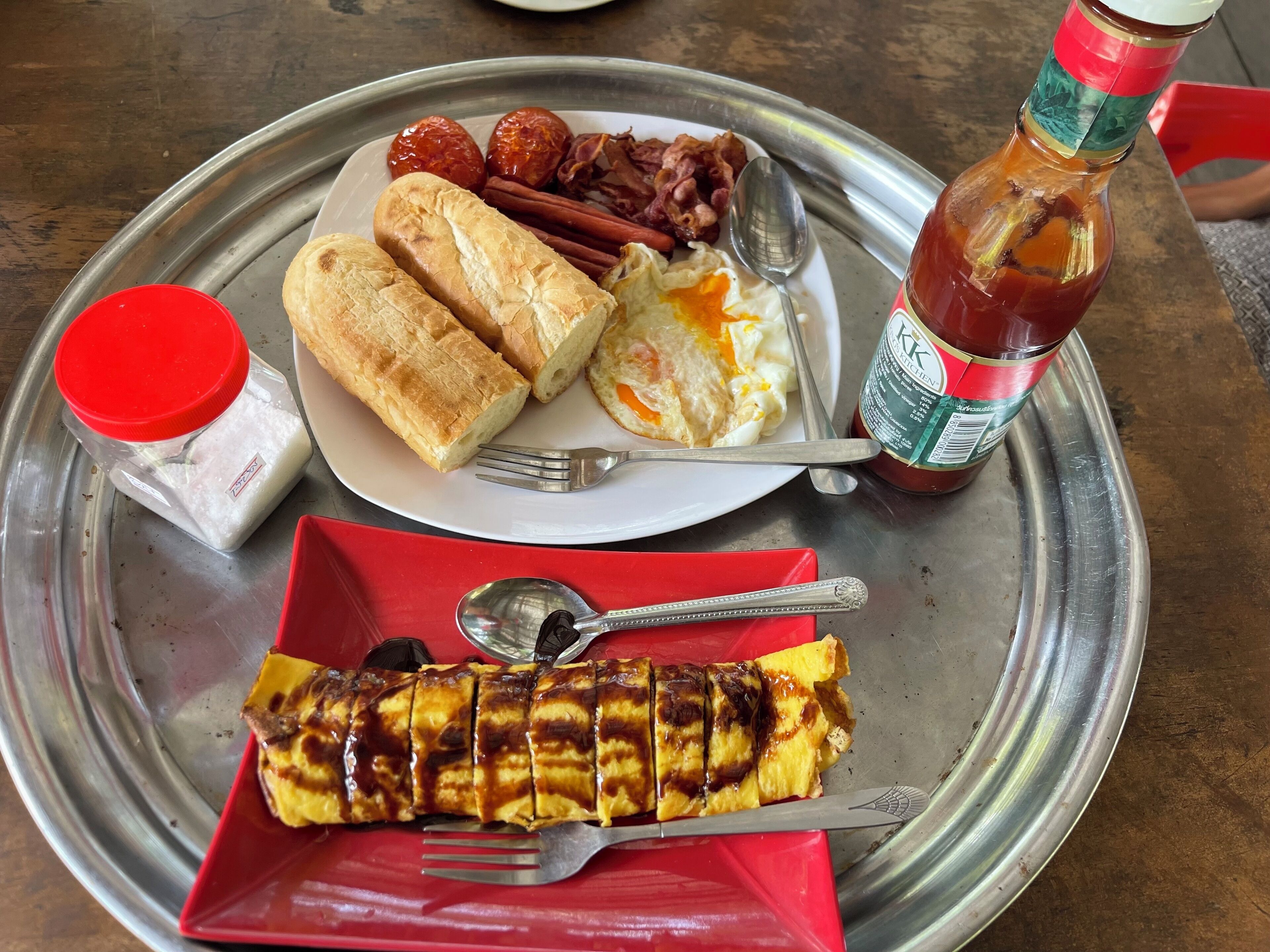 A meal tray with bread, eggs, bacon, sausages, and a bottle of ketchup.