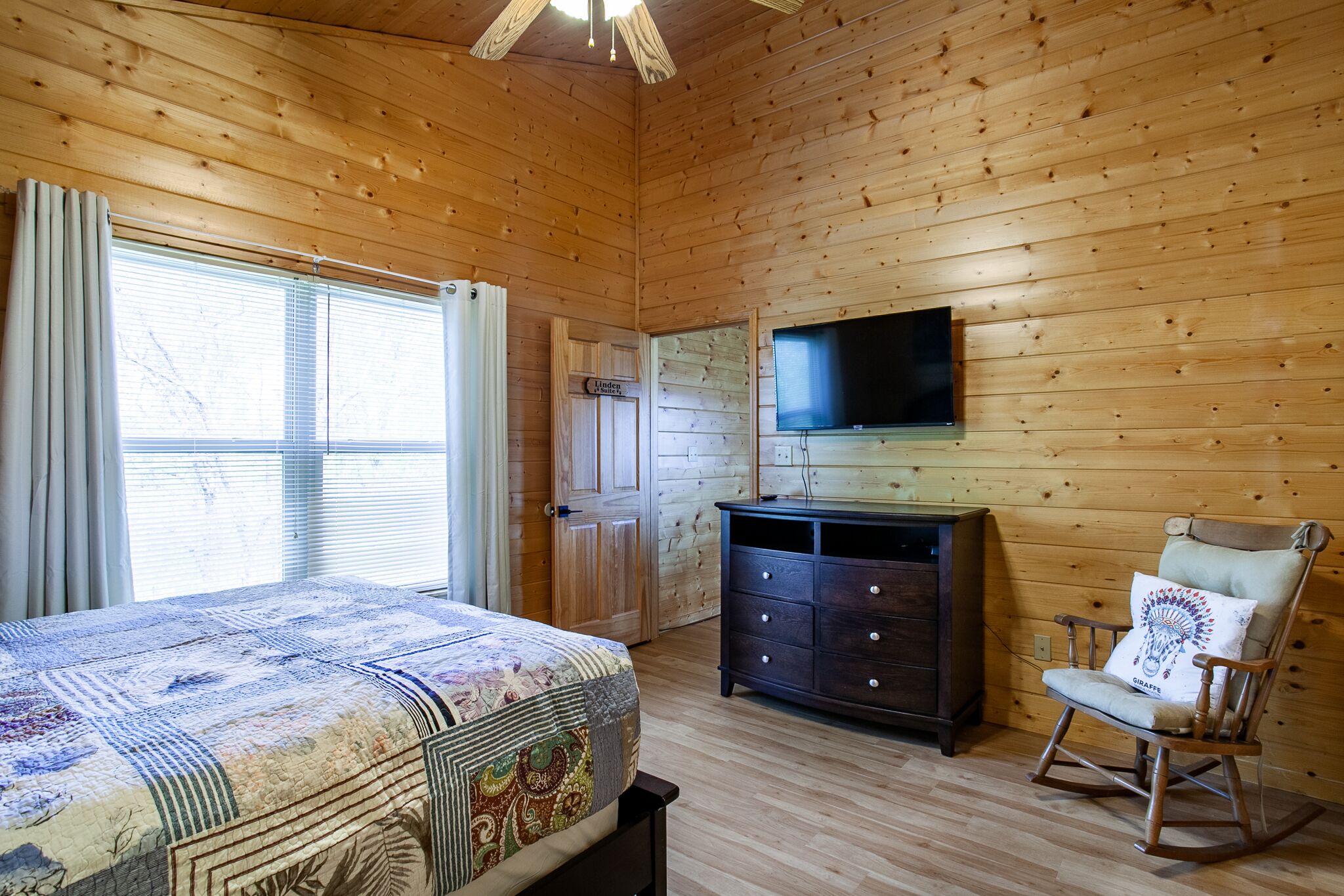 A wooden-paneled room with a bed, a dresser, a rocking chair, and a TV mounted on the wall.