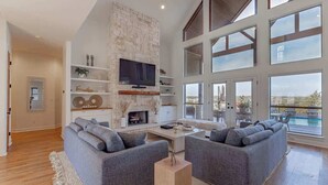 A spacious living room with a stone fireplace, grey sofas, a flat-screen TV, and large windows offering a view of the outdoors. - Sleeps 20 | Pool, Game Room | Luxury Fall Retreat (Dripping Springs)