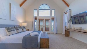 A bedroom with a large bed, a wooden bench, a TV, and a view of the outdoors through a door. - Sleeps 20 | Pool, Game Room | Luxury Fall Retreat (Dripping Springs)