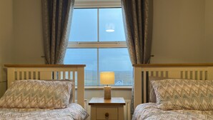 A room with two beds, each with patterned bedding, a bedside table with a lamp, and a window with curtains. - Cottage 427 - Clifden - sleeps 6 guests  in 3 bedrooms (Clifden)