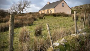 A house with a sloped roof, surrounded by a grassy field and a wooden fence. - Cottage 427 - Clifden - sleeps 6 guests  in 3 bedrooms (Clifden)