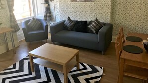 Sala de estar com sofá cinza, duas poltronas, mesa de centro de madeira e mesa de jantar com cadeiras. Apresenta papel de parede estampado e um quadro na parede.