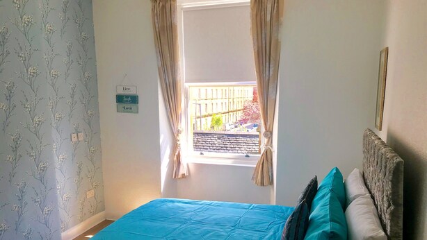 Quarto com colcha azul, cabeceira de madeira e janela com cortinas.