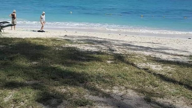 Ein Strand mit zwei Personen, die im Sand stehen, einem Segelboot in der Ferne und einem klaren, blauen Himmel.