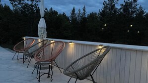 Un espace extérieur aménagé avec des chaises en osier et un parasol blanc, entouré d’une terrasse en bois et d’arbres.
