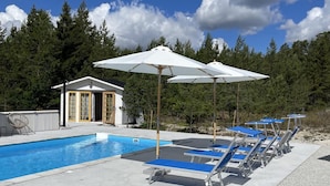 Une piscine avec des chaises longues et des parasols, un petit bâtiment aux portes vitrées et un paysage boisé en arrière-plan.