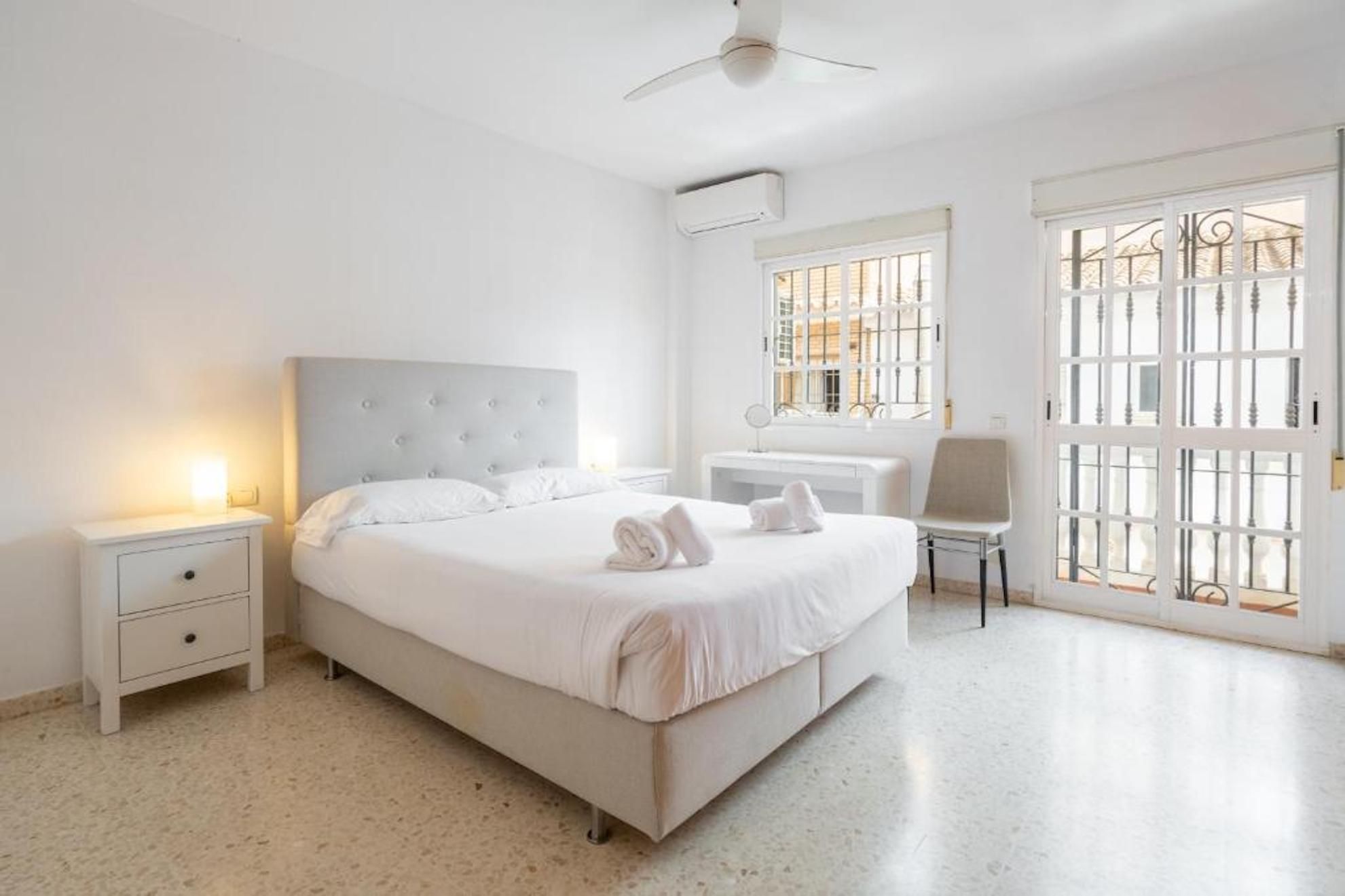 A bedroom with a bed, bedside tables, a chair, a window with metal bars, and an air conditioning unit.