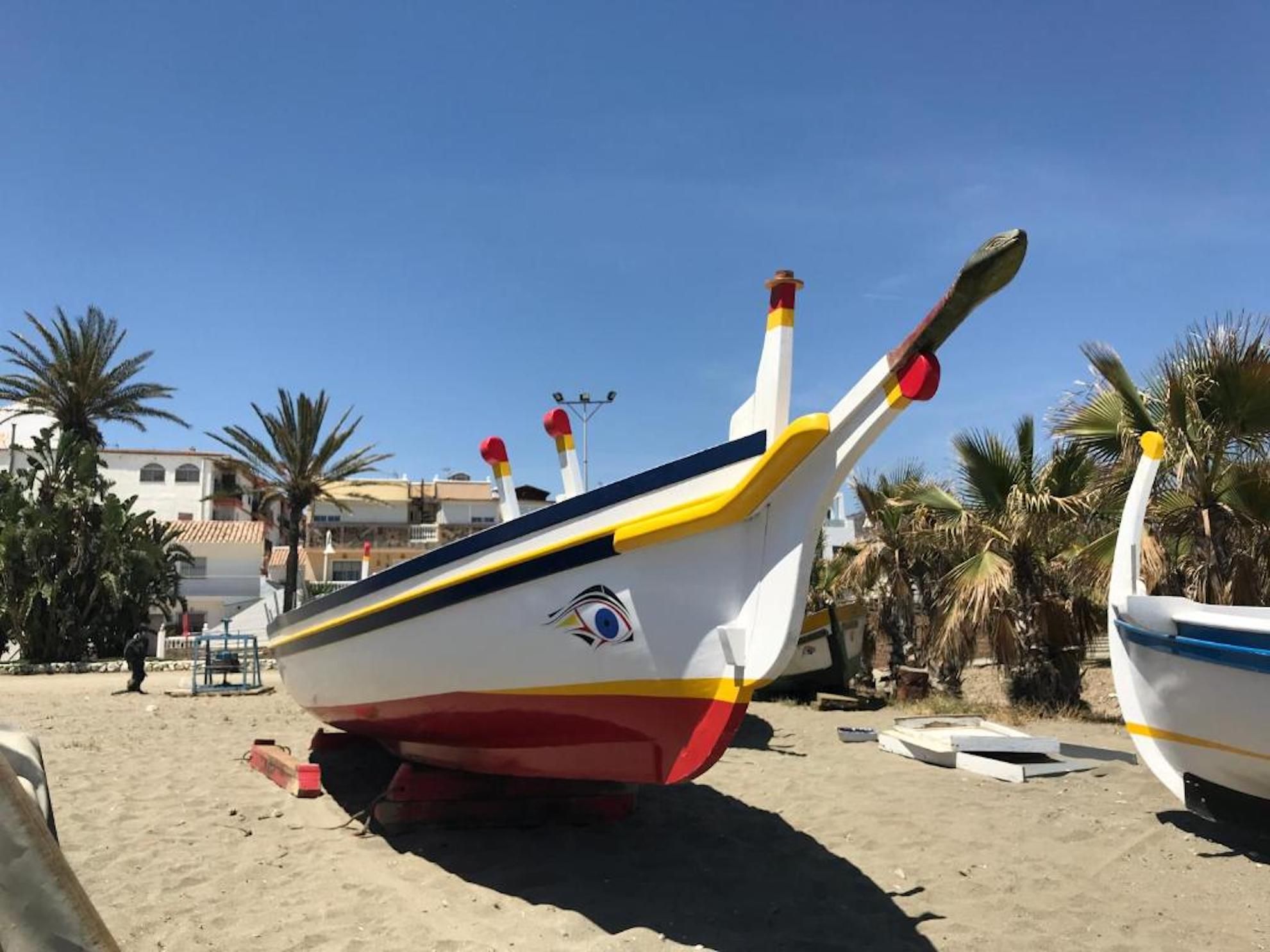 Un bote con un logotipo distintivo, descansando sobre una playa arenosa con palmeras y edificios al fondo.